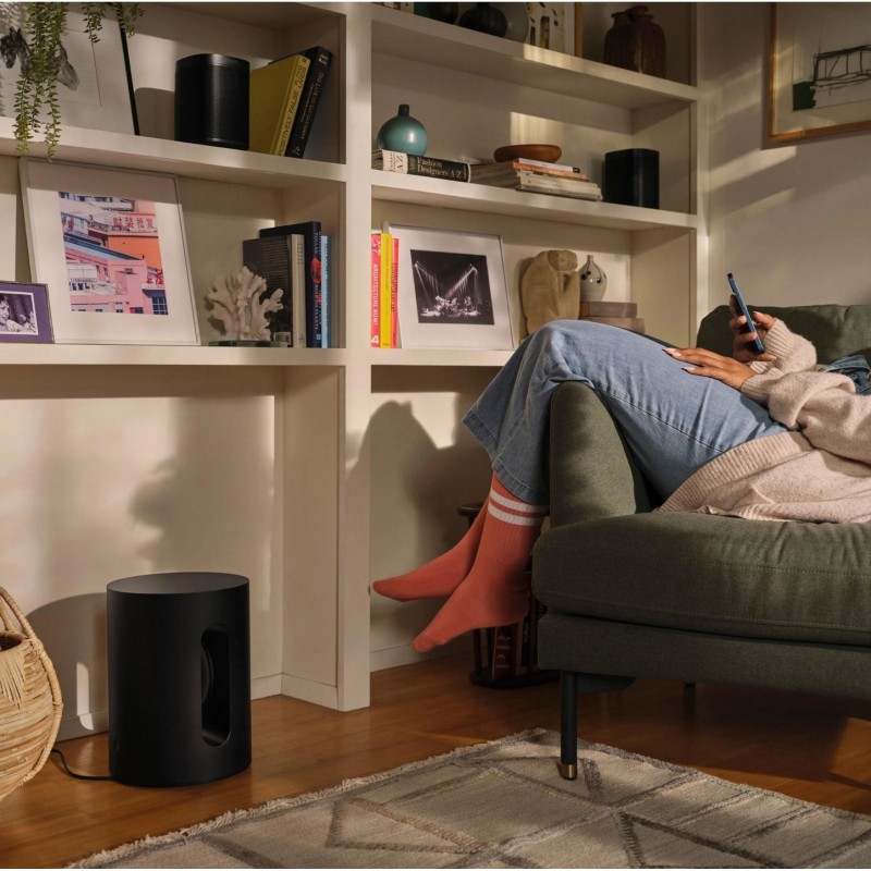 Sonos Bundle Beam Mini en negro, el Sub Mini en el suelo junto a una estantería y un sofá con una persona mirando el móvil